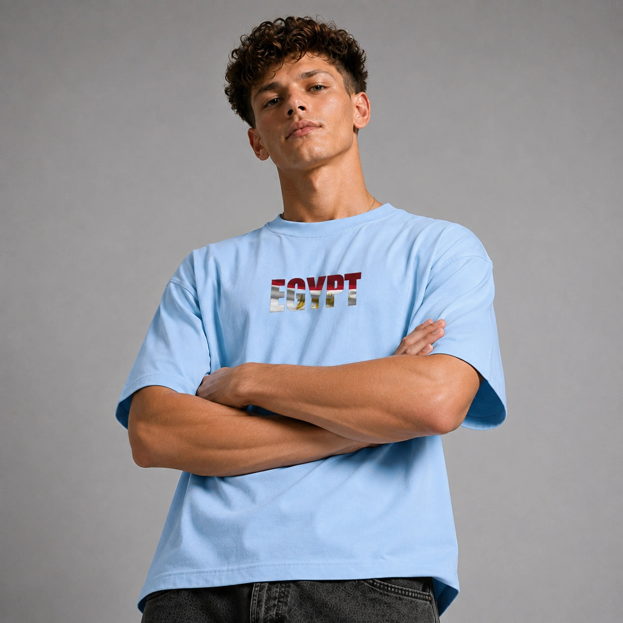 A young man with curly brown hair wearing a light blue t-shirt with 'EGYPT' printed across the chest in the colors of the Egyptian flag, posed against a neutral gray studio background.