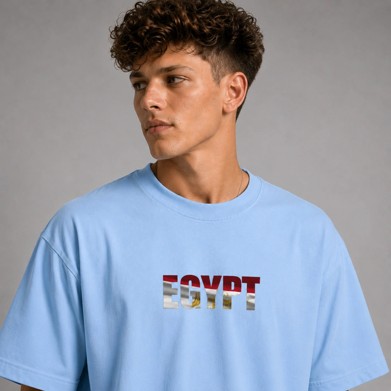 A man wearing a light blue oversized t-shirt with 'EGYPT' printed across the chest in the style of the American flag, set against a plain gray studio background.