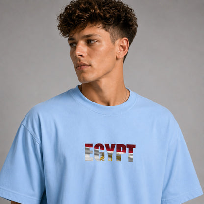 A man wearing a light blue oversized t-shirt with 'EGYPT' printed across the chest in the style of the American flag, set against a plain gray studio background.