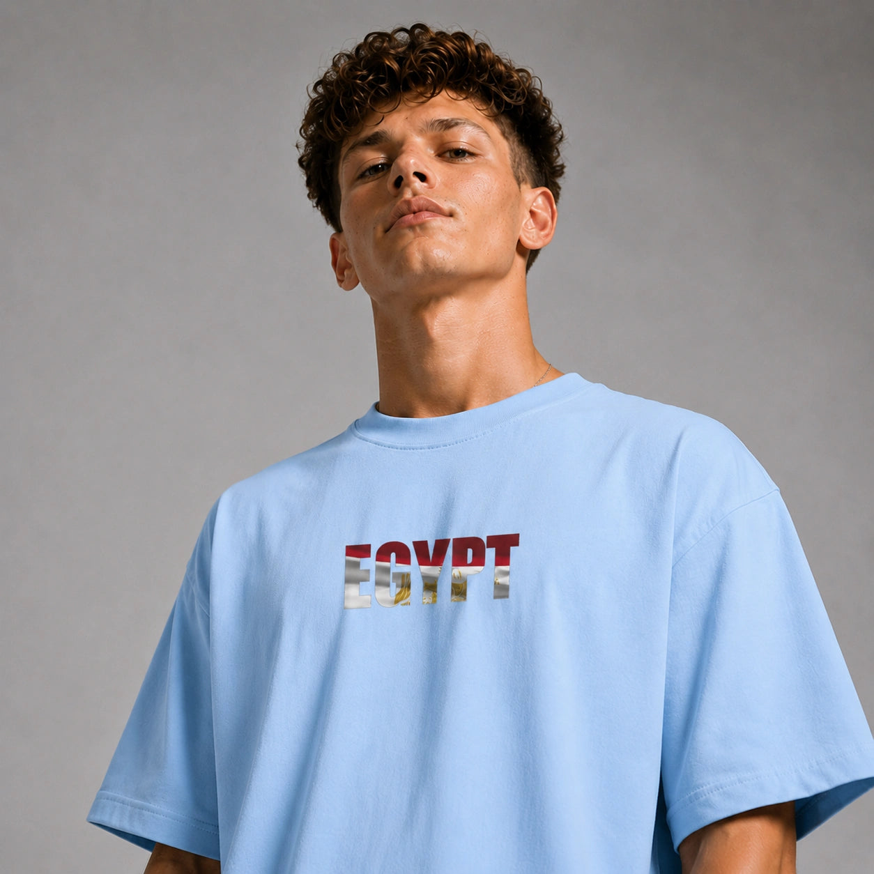 A young man with curly brown hair wearing a light blue t-shirt with 'EGYPT' printed across the chest in the style of the Egyptian flag, set against a plain gray studio background.