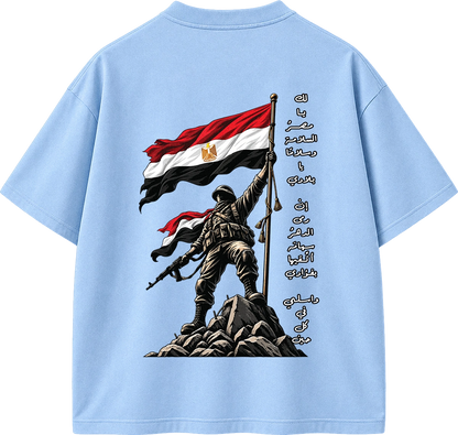 A light blue short-sleeve t-shirt shown from the back, featuring a large graphic print of a soldier holding an Egyptian flag on a rocky outcrop, with vertical Arabic text printed alongside.