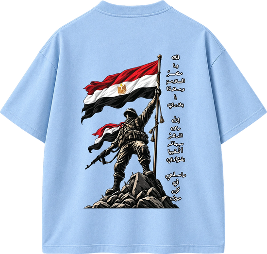 A light blue short-sleeve t-shirt shown from the back, featuring a large graphic print of a soldier holding an Egyptian flag on a rocky outcrop, with vertical Arabic text printed alongside.
