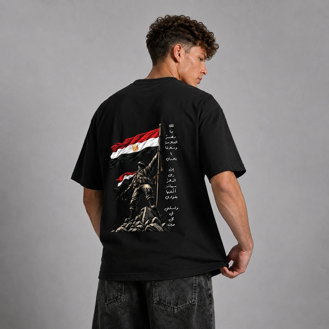 A man wearing a black t-shirt with a large back graphic of the Egyptian flag, a soldier statue, and Arabic text, shown from behind against a gray studio background.
