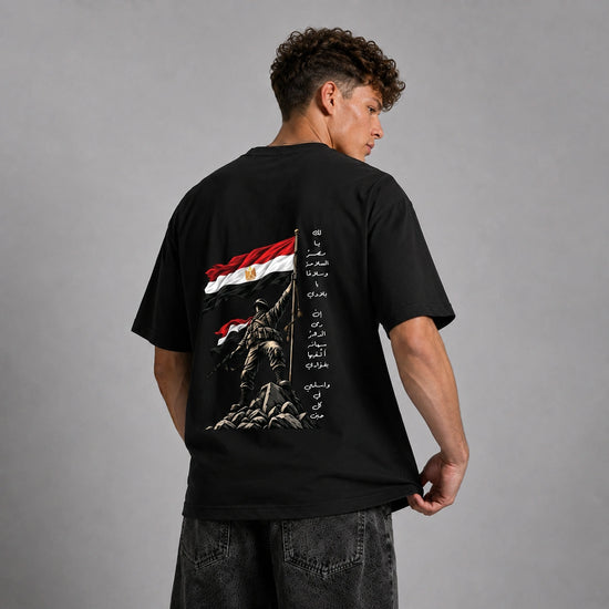 A man wearing a black t-shirt with a large back graphic of the Egyptian flag, a soldier statue, and Arabic text, shown from behind against a gray studio background.
