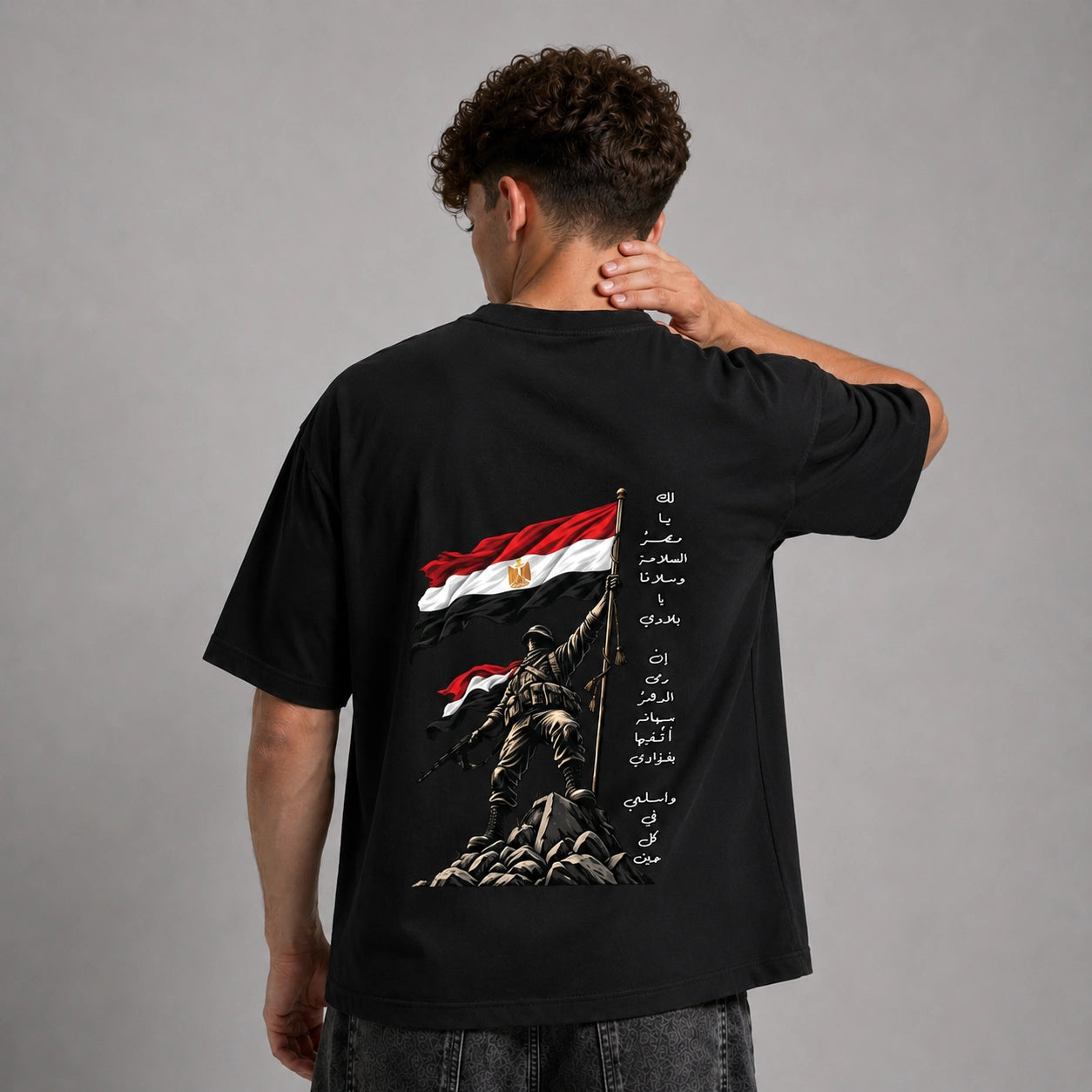 A man viewed from behind wearing a black graphic t-shirt with a large Egyptian flag and soldier design on the back, set against a plain gray studio background.
