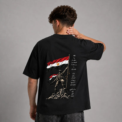 A man viewed from behind wearing a black graphic t-shirt with a large Egyptian flag and soldier design on the back, set against a plain gray studio background.