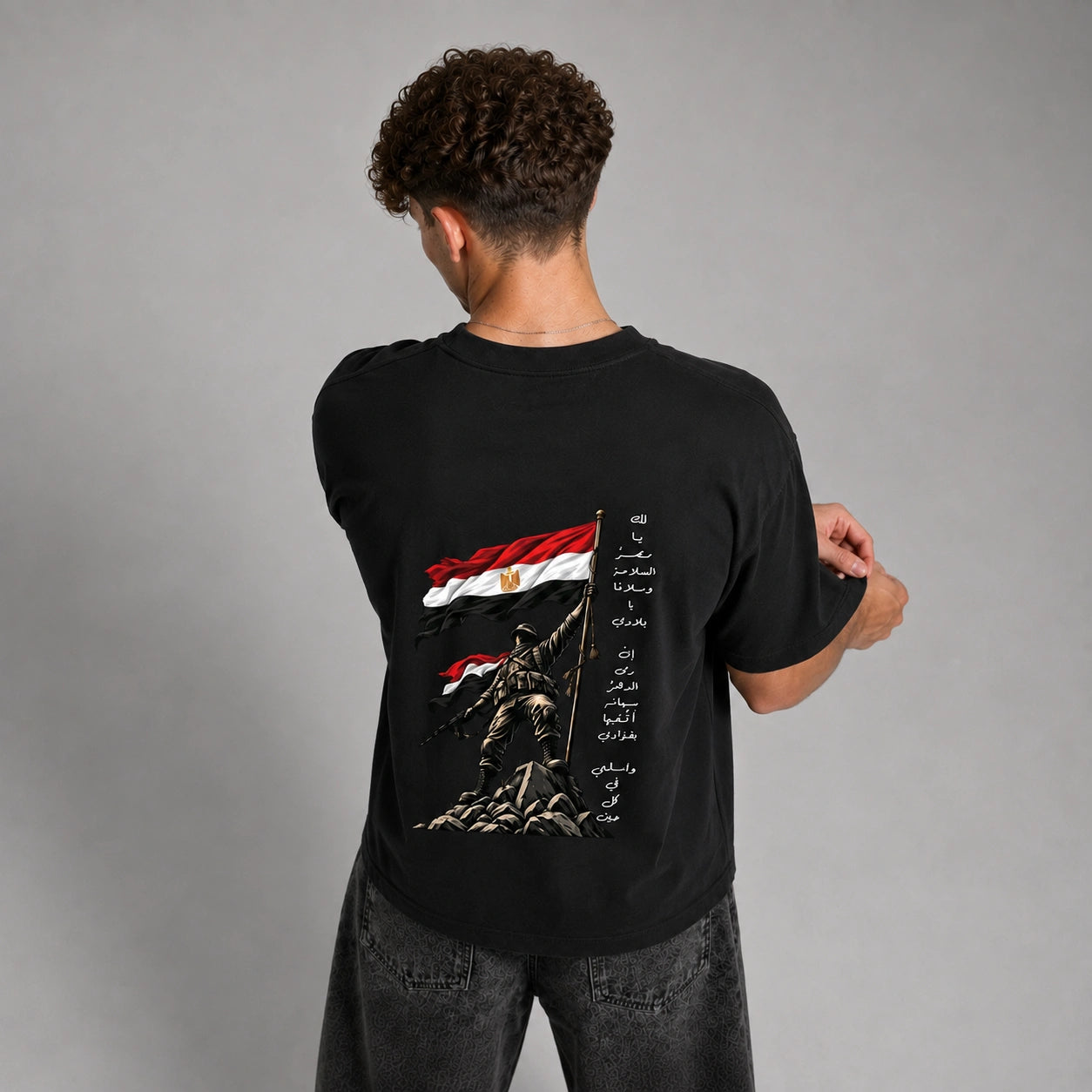 A man viewed from behind wearing a black t-shirt with a large graphic print of the Egyptian flag, a soldier holding a flagpole, and Arabic calligraphy on the back, paired with dark gray jeans against a gray studio background.