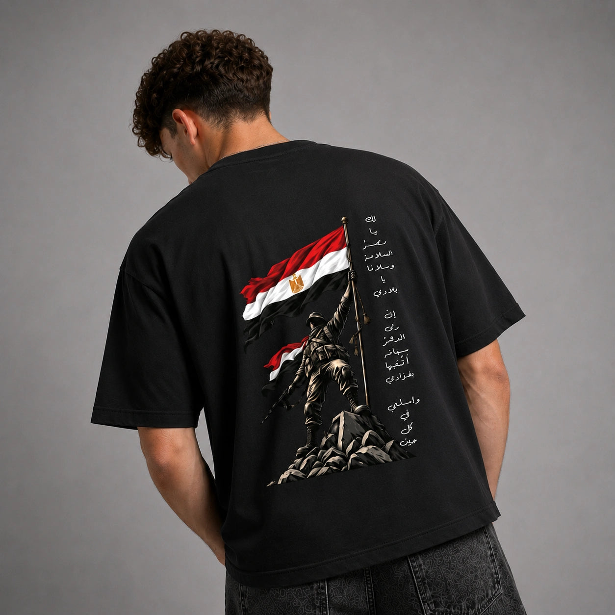 A man viewed from behind wearing a black t-shirt with a large graphic print on the back. The print features the Egyptian flag, a soldier statue, and vertical Arabic text.