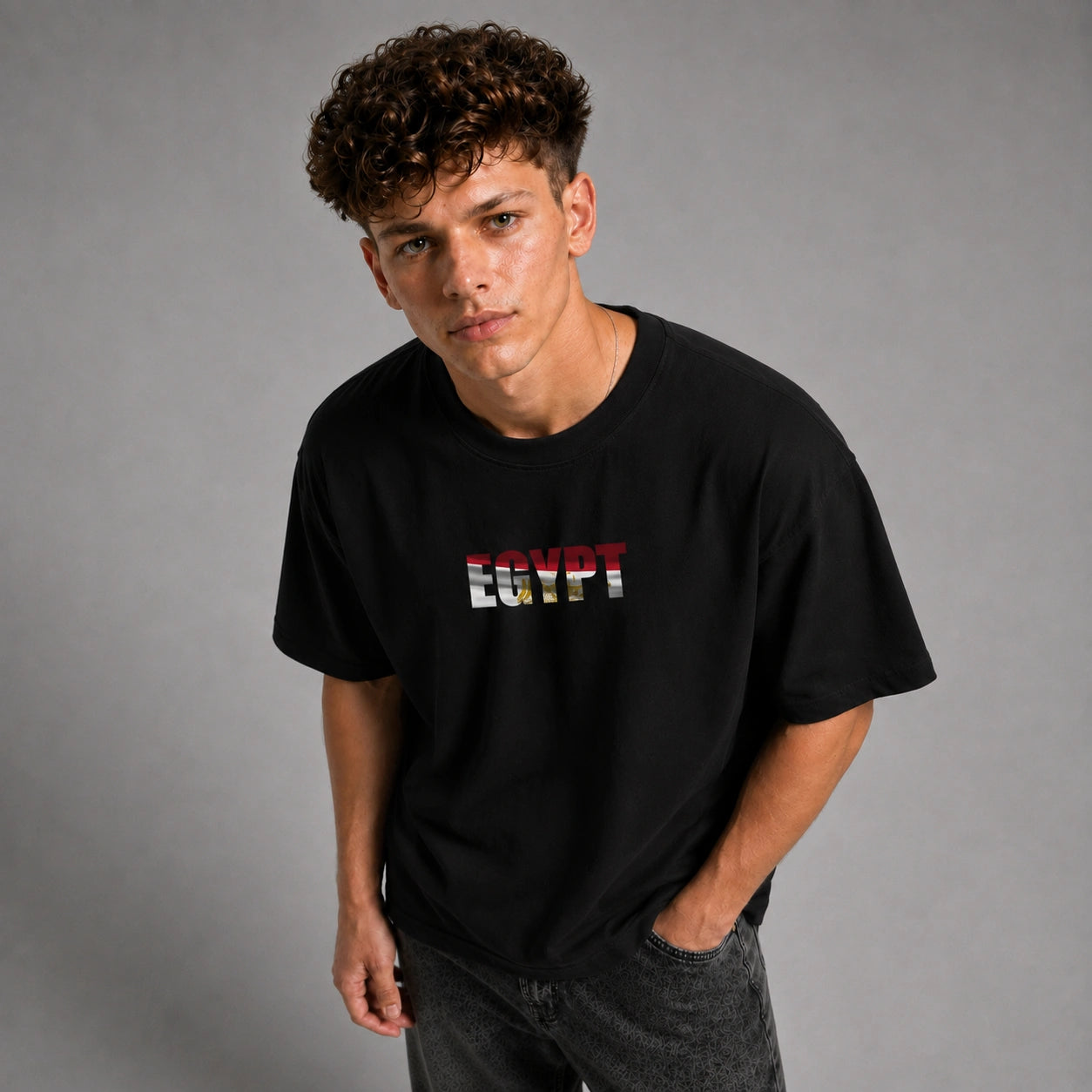 A young man with curly brown hair wearing a black t-shirt with 'EGYPT' printed across the chest in the colors of the Egyptian flag, paired with dark gray jeans, against a solid gray studio background.