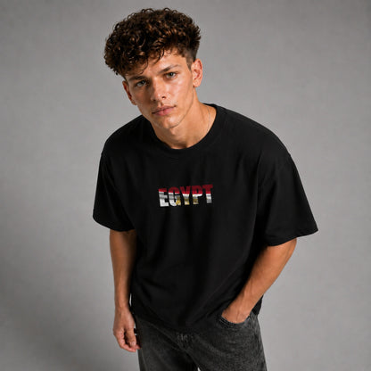 A young man with curly brown hair wearing a black t-shirt with 'EGYPT' printed across the chest in the colors of the Egyptian flag, paired with dark gray jeans, against a solid gray studio background.