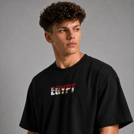 A man wearing a black short-sleeve t-shirt with 'EGYPT' printed across the chest in the style of the Egyptian flag, set against a plain gray studio background.