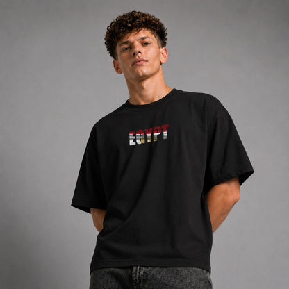 A young man wearing a black t-shirt with 'EGYPT' printed across the chest in the colors of the Egyptian flag, posed against a neutral gray studio background.