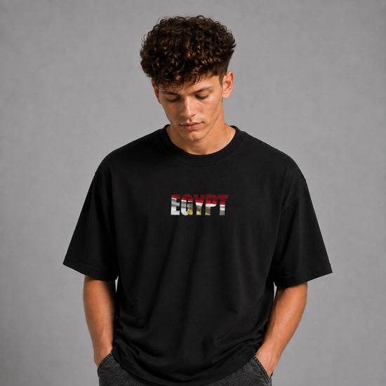 A man wearing a black crewneck t-shirt with 'EGYPT' printed across the chest in the style of the Egyptian flag, standing against a plain gray studio background.