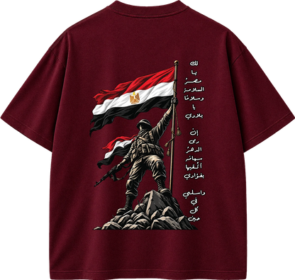 A dark maroon short-sleeve t-shirt shown from the back, featuring a graphic print of a soldier holding the Egyptian flag on a rocky outcrop, with vertical Arabic text printed on the right side.