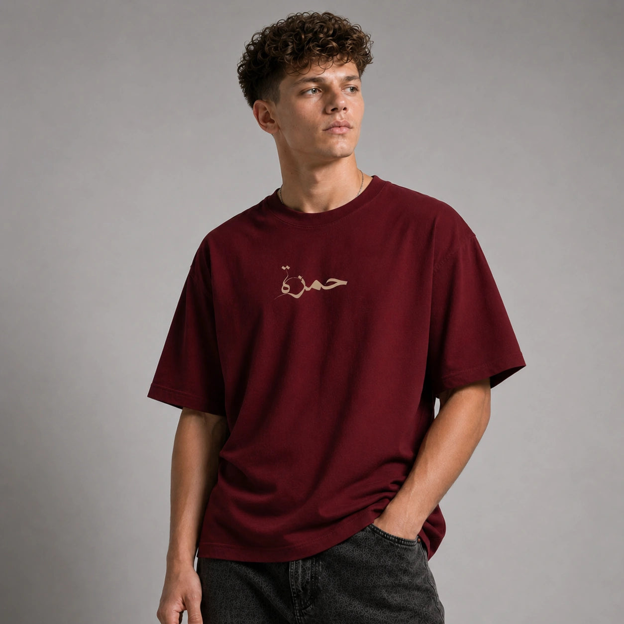 A young man with curly brown hair wearing an oversized maroon t-shirt with gold Arabic calligraphy on the chest, paired with dark gray jeans, standing against a neutral gray studio background.