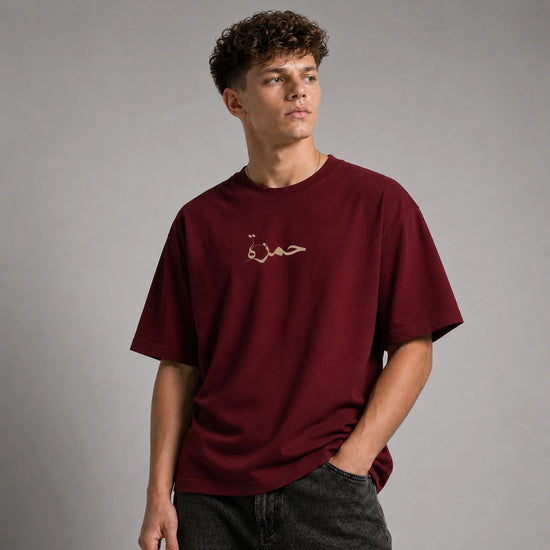 A young man with curly brown hair wearing an oversized maroon t-shirt with gold Arabic calligraphy on the chest, paired with dark gray jeans, standing against a neutral gray studio background.