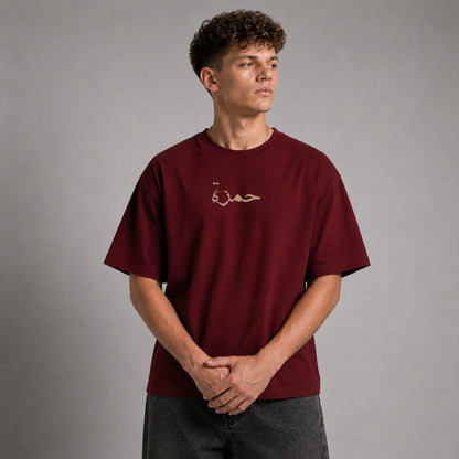 A man wearing a maroon short-sleeve t-shirt with a small beige Arabic calligraphy graphic on the left chest, standing against a neutral gray studio background.