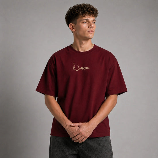 A man wearing a maroon short-sleeve t-shirt with a small beige Arabic calligraphy graphic on the left chest, standing against a neutral gray studio background.