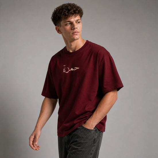 A man wearing a maroon crewneck t-shirt with gold Arabic calligraphy on the chest, paired with dark gray jeans, photographed against a neutral gray studio background.