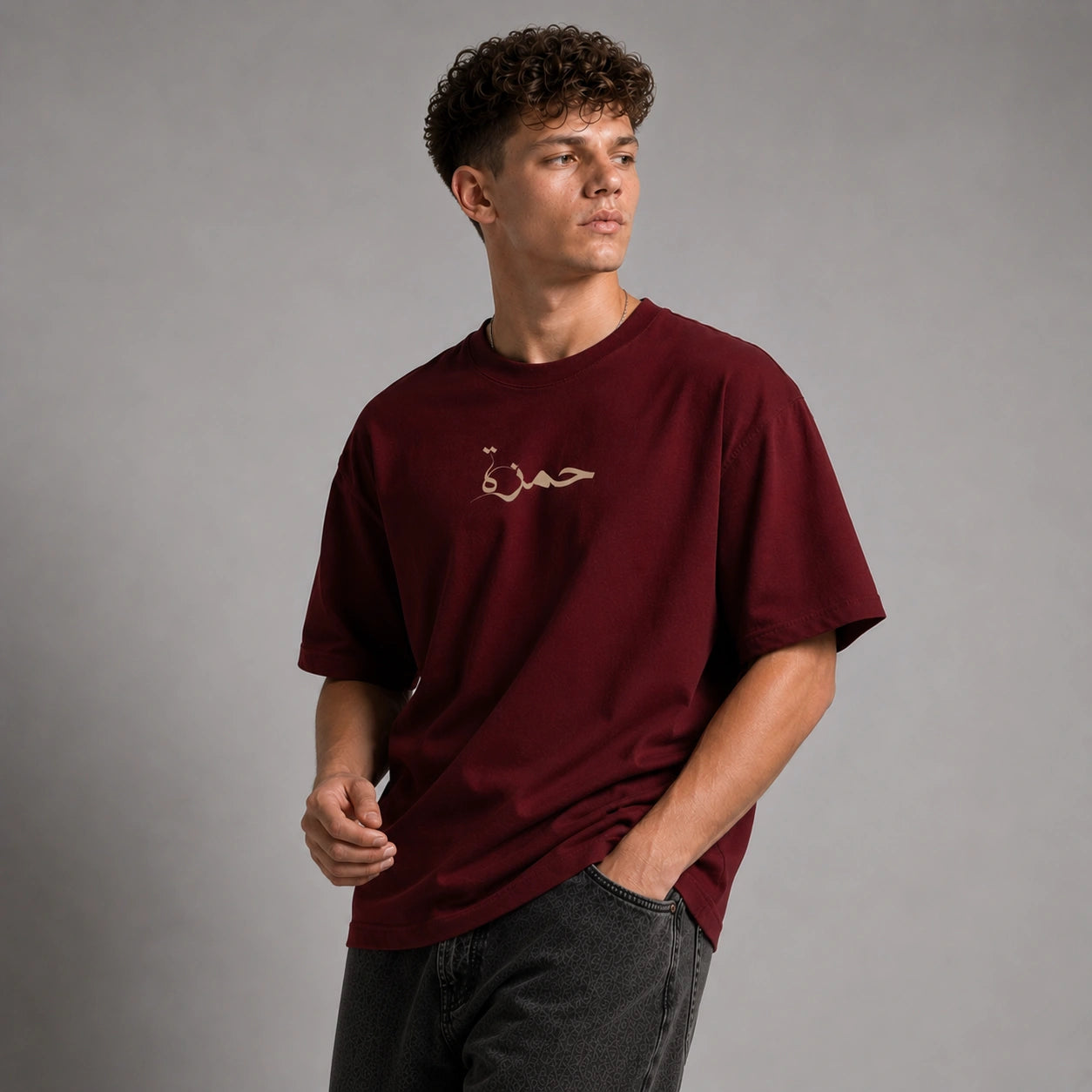 A man wearing a maroon short-sleeve t-shirt with a small gold embroidered Arabic script on the chest, paired with dark gray jeans, set against a plain gray studio background.