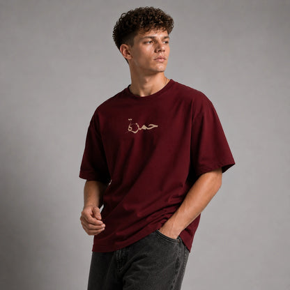 A man wearing a maroon short-sleeve t-shirt with a small gold embroidered Arabic script on the chest, paired with dark gray jeans, set against a plain gray studio background.