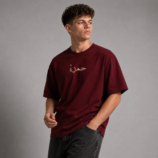 A man wearing a maroon short-sleeve t-shirt with a small gold embroidered Arabic script on the chest, paired with dark gray jeans, set against a plain gray studio background.