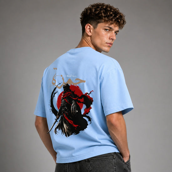 A man wearing a light blue t-shirt with a large graphic print on the back, photographed from behind against a neutral gray studio background.