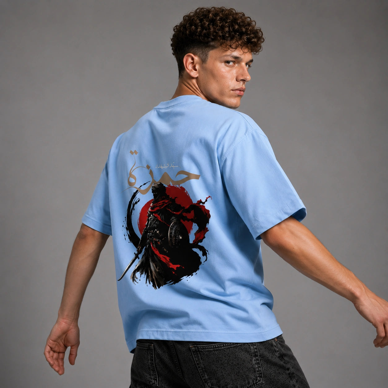 A man wearing a light blue t-shirt with a large graphic print on the back, featuring a samurai warrior and a red sun, photographed against a neutral gray studio background.