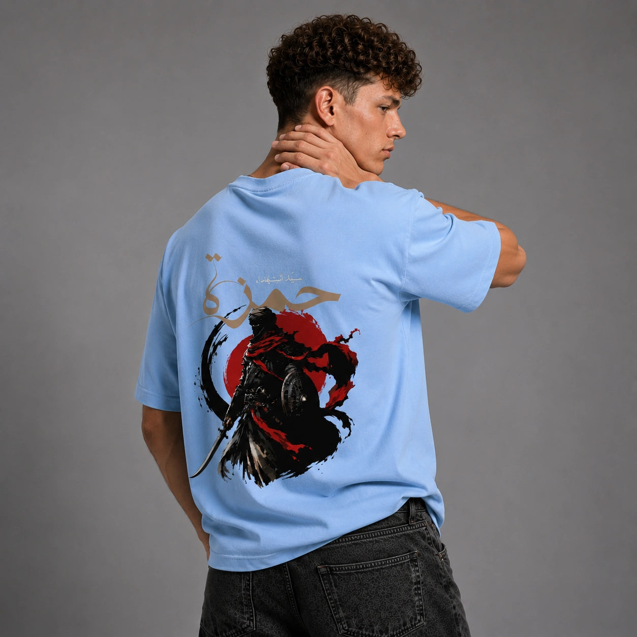 A man wearing a light blue t-shirt with a large graphic print on the back, shown from behind against a gray studio background.