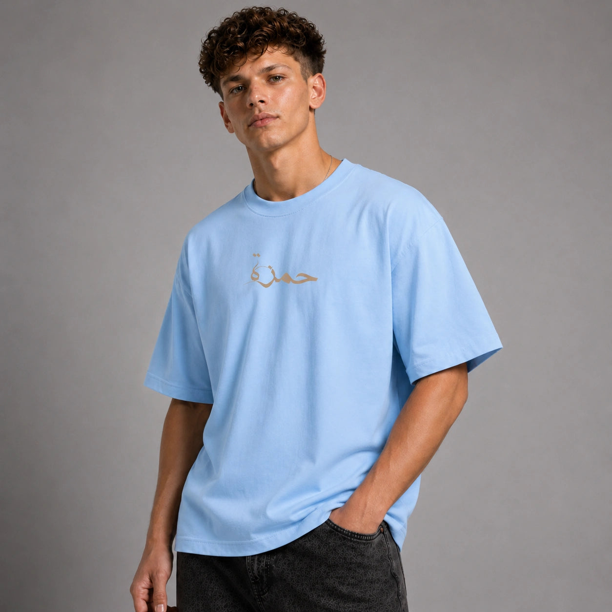 A young man wearing a light blue crewneck t-shirt with gold Arabic calligraphy on the chest, paired with dark gray jeans, set against a solid gray studio background.