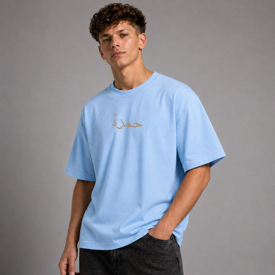 A young man wearing a light blue crewneck t-shirt with gold Arabic calligraphy on the chest, paired with dark gray jeans, set against a solid gray studio background.