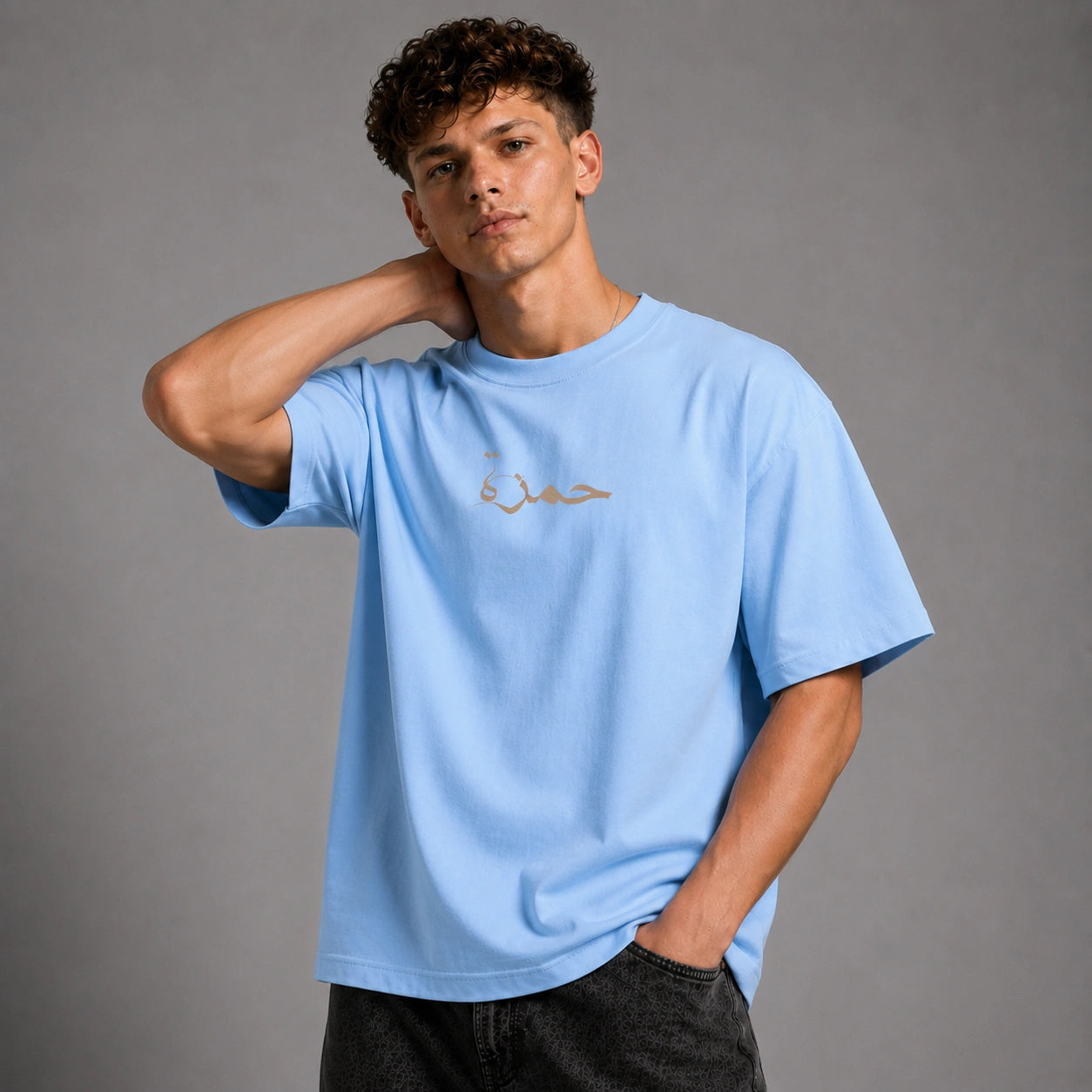 A young man with curly brown hair wearing a light blue crewneck t-shirt with gold Arabic calligraphy on the chest, paired with dark gray jeans, posing against a solid gray studio background.