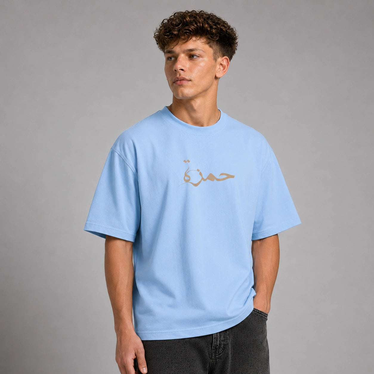 A young man with curly brown hair wearing an oversized light blue t-shirt with a gold Arabic calligraphy graphic on the chest, paired with dark gray jeans, photographed against a neutral gray studio background.