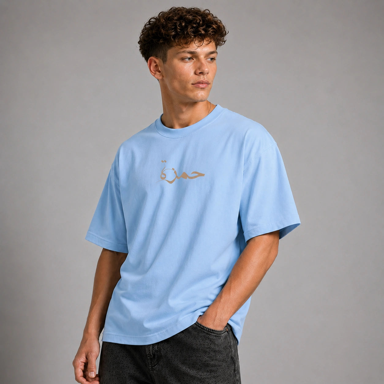 A young man with curly brown hair wearing a light blue crewneck t-shirt with a small gold Arabic calligraphy graphic on the chest, paired with dark gray jeans, standing against a solid gray studio background.