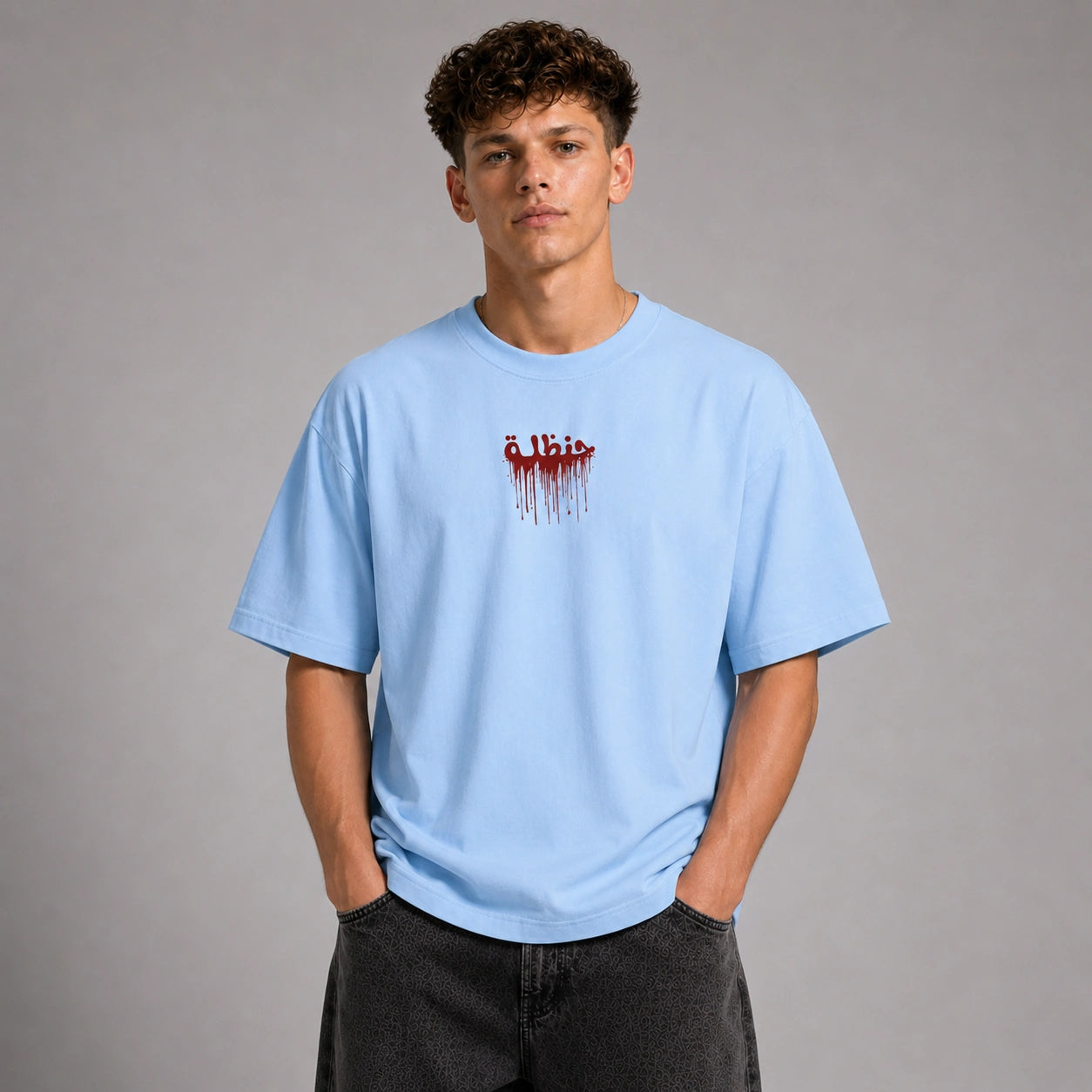 A young man wearing a light blue crewneck t-shirt with a red dripping 'Elbist' logo on the chest, paired with dark gray jeans, standing against a neutral gray studio background.