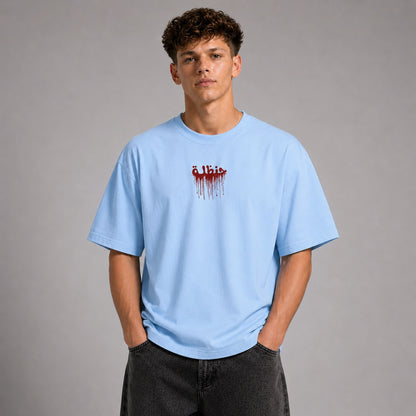 A young man wearing a light blue crewneck t-shirt with a red dripping 'Elbist' logo on the chest, paired with dark gray jeans, standing against a neutral gray studio background.