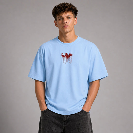 A young man wearing a light blue crewneck t-shirt with a red dripping 'Elbist' logo on the chest, paired with dark gray jeans, standing against a neutral gray studio background.