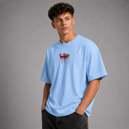 A young man wearing a light blue crewneck t-shirt with a red dripping graphic on the chest, paired with dark gray jeans, standing against a neutral gray studio background.