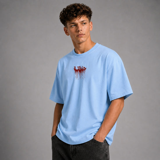 A young man wearing a light blue crewneck t-shirt with a red dripping graphic on the chest, paired with dark gray jeans, standing against a neutral gray studio background.