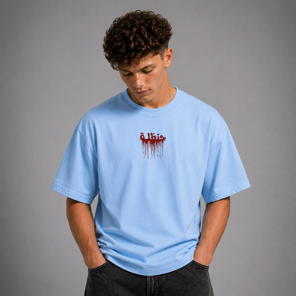 A young man with curly dark hair wearing a light blue t-shirt with a red dripping text graphic on the chest, posed against a solid gray studio background.