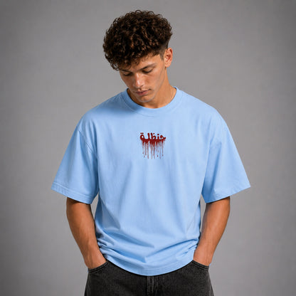 A young man with curly dark hair wearing a light blue t-shirt with a red dripping text graphic on the chest, posed against a solid gray studio background.