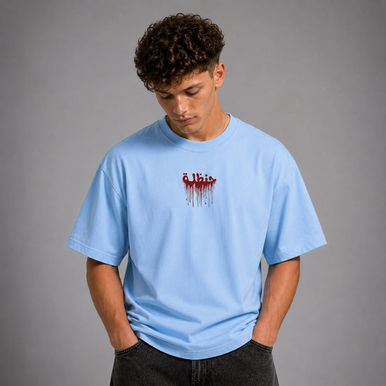 A young man with curly dark hair wearing a light blue t-shirt with a red dripping text graphic on the chest, posed against a solid gray studio background.