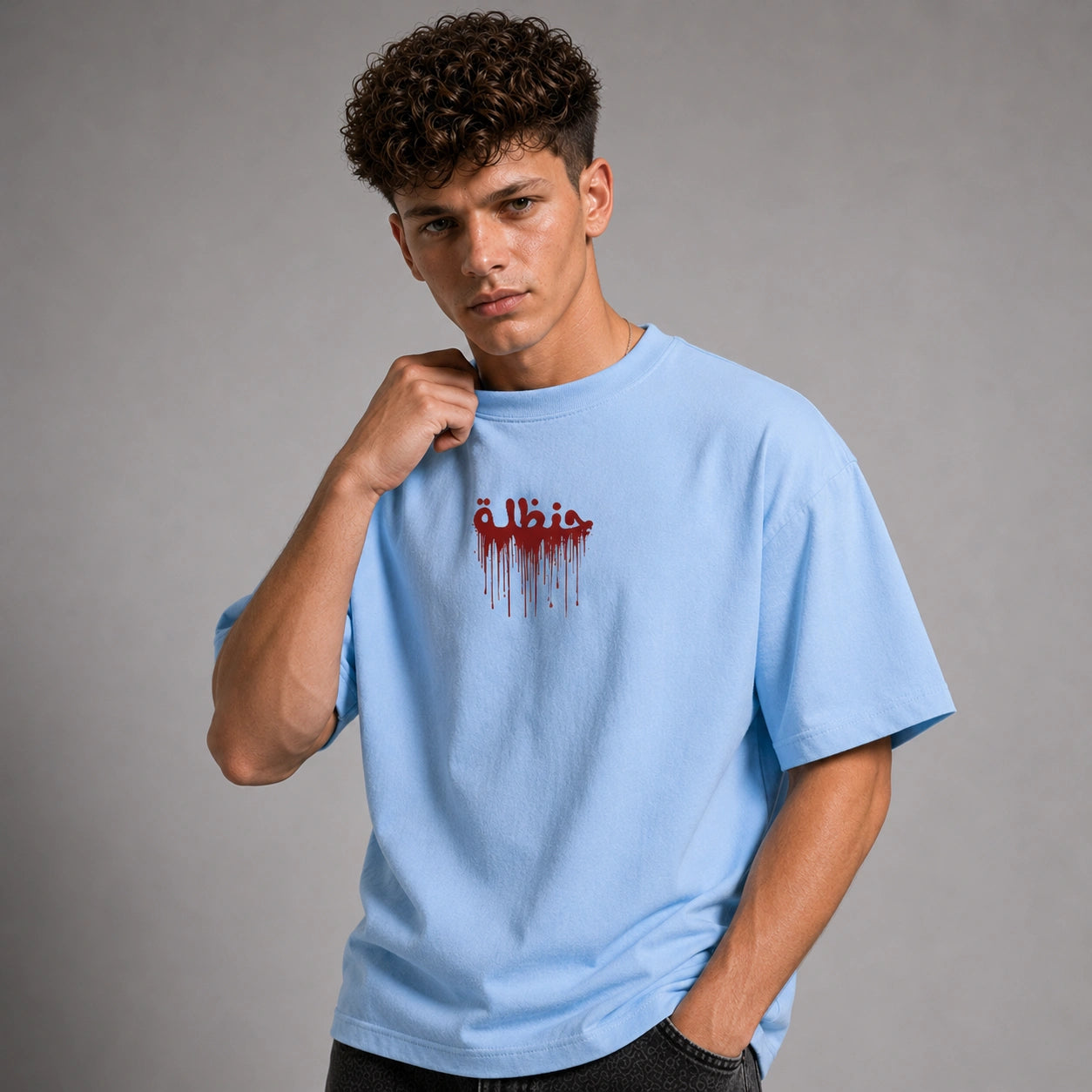A young man with curly brown hair wearing a light blue t-shirt with a red dripping Arabic text graphic on the chest, posed against a neutral gray studio background.