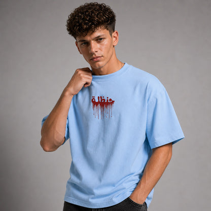 A young man with curly brown hair wearing a light blue t-shirt with a red dripping Arabic text graphic on the chest, posed against a neutral gray studio background.