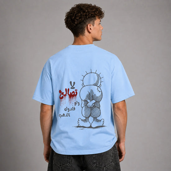 A man shown from the back wearing a light blue t-shirt with a large graphic print on the back. The graphic features a stylized cartoon character in black and gray, red Arabic text, and black Arabic calligraphy.