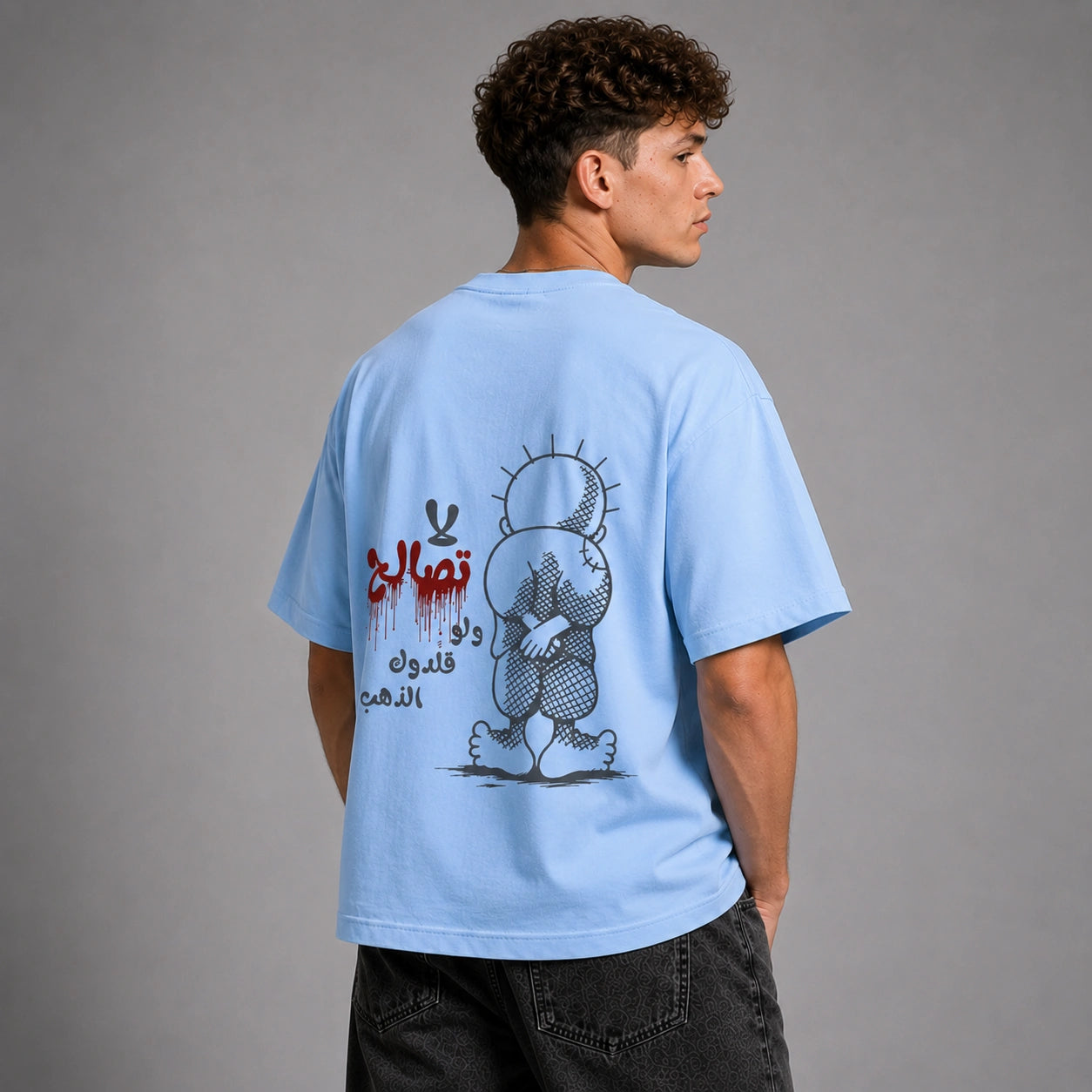 A man wearing a light blue t-shirt, shown from the back. The shirt features a graphic print of a cartoon bear with Arabic text in red and black.