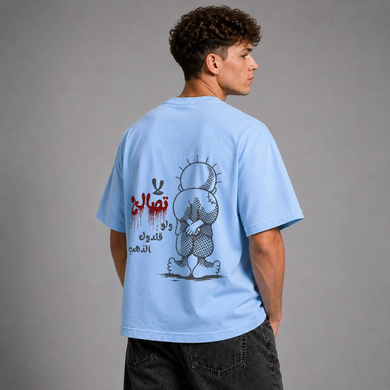 A man wearing a light blue t-shirt, shown from the back. The shirt features a graphic print of a cartoon bear with Arabic text in red and black.