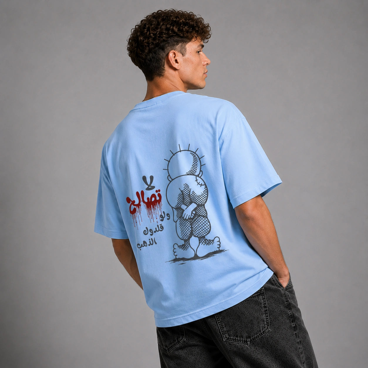 A man wearing a light blue graphic t-shirt, viewed from behind to show the large back print. The print features a black and white cartoon character and Arabic text in black and red.