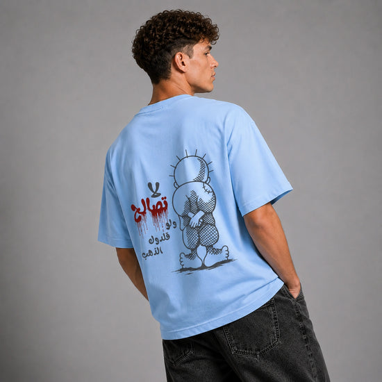 A man wearing a light blue graphic t-shirt, viewed from behind to show the large back print. The print features a black and white cartoon character and Arabic text in black and red.