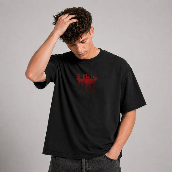 A man wearing a black crewneck t-shirt with a red dripping 'Elvis' graphic on the chest, posed against a neutral gray background.